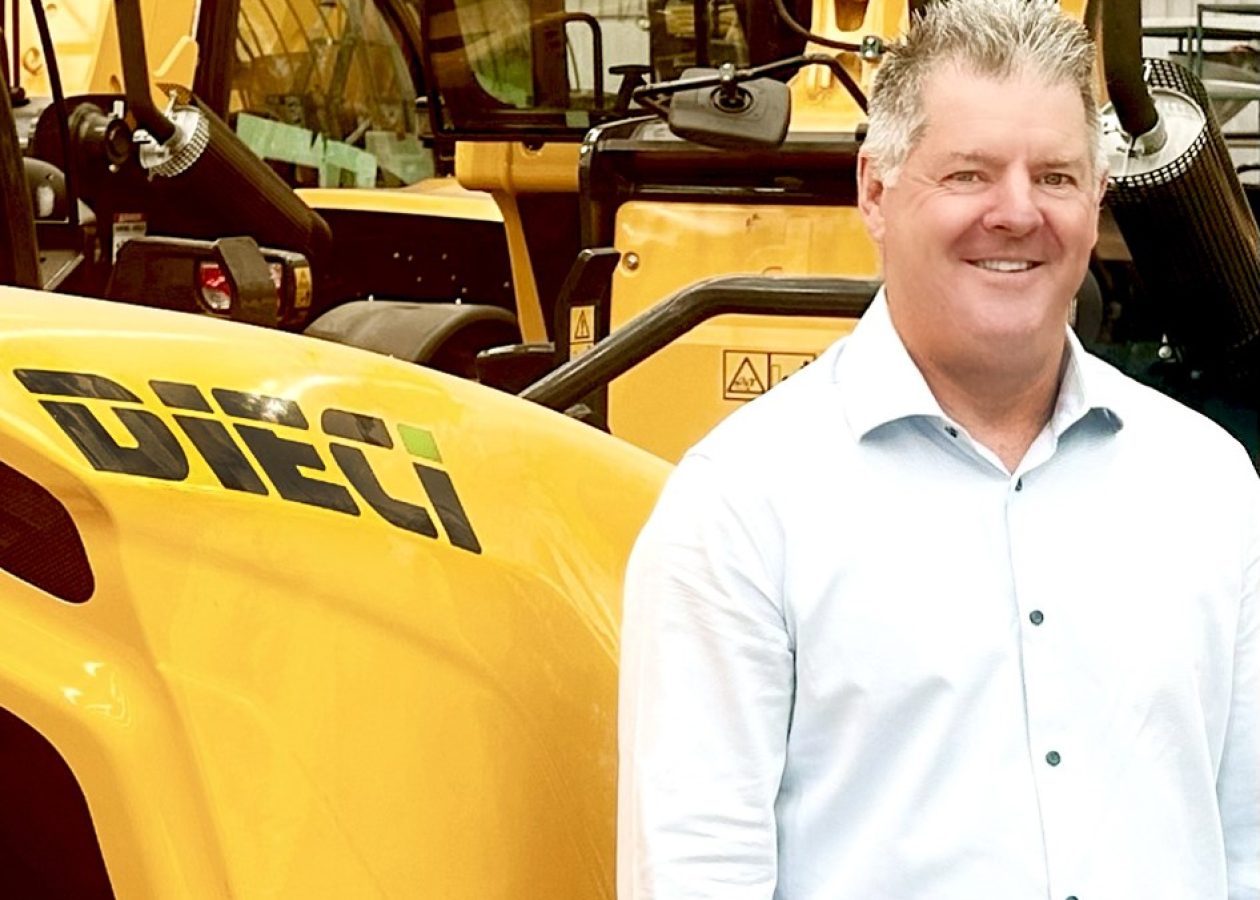 Smiling man in a light blue shirt stands beside a yellow Dieci construction machine.
